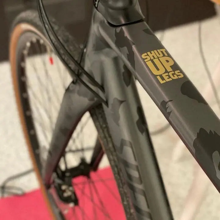 Load image into Gallery viewer, Gold 'SHUT UP LEGS' vinyl sticker on a gray camo bike top tube (set of 2)

